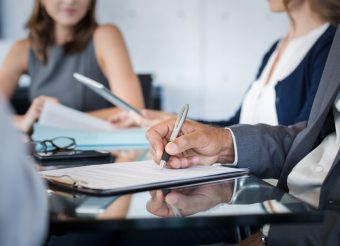 Closeup of hand of businessman holding pen and analyzing report during board meeting. Business people writing on document during annual meeting. Detail hand of business man fill the business contract.